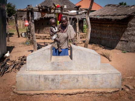 An older African woman holding an infant stands by a concrete grave next to some huts 
