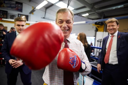 Farage buzëqesh ndërsa bën sikur godet fotografin me doreza të kuqe boksi në një ring boksi.