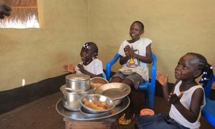 Three children clap their hands while they sit around a small table with dishes balanced on it.