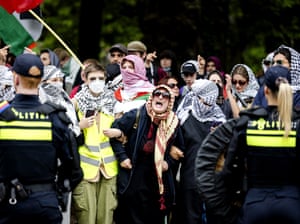 Protesters wearing keffiyehs and holding Palestinian flags face police officers in dark uniforms marked “Politie” during a demonstration