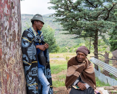 Two men wearing hats and blankets wait outside a wooden home