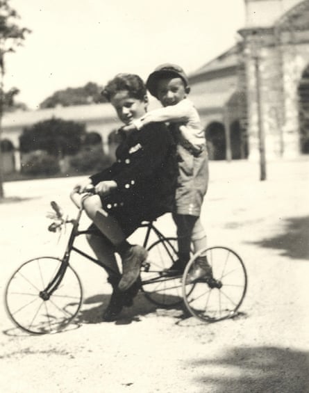 A black-and-white photograph of two boys on a tricycle in front of a building, one sitting on the seat wearing a dark velvet short suit, and the other standing on the back wearing dungarees and a flat cap.
