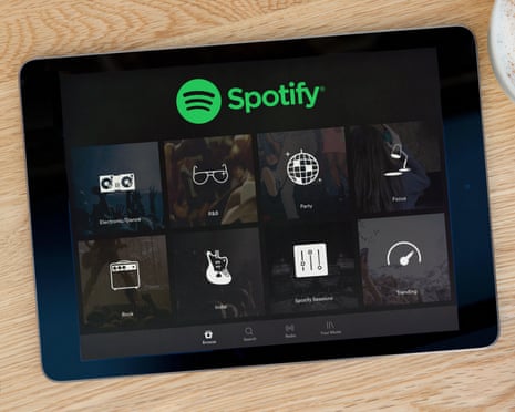 A tablet displaying the Spotify app with music genre categories sits on a wooden table beside a coffee cup