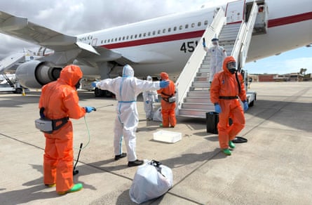A person in a white hazmat suit and visor stands on the tarmac next to an aircraft and is sprayed by a person wearing an orange protective suit and visor while others in protective clothing look on.