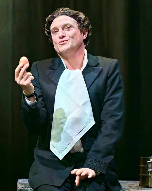 Mathew Horne as Malvolio, in a tuxedo with an untucked napkin, smiles while holding an egg on stage