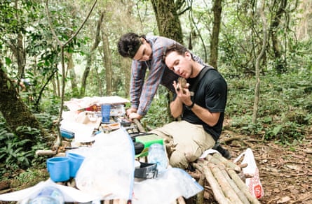 Em uma clareira na floresta, um homem se inclina sobre uma longa mesa coberta de apetrechos científicos, olhando por cima do ombro de um segundo homem que está sentado examinando as asas de um pequeno pássaro que ele está segurando.