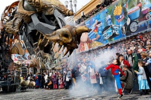 A young boy dressed in a Spider Man costume stands in front of a crowd as large model of a fire-breathing dragon snorts smoke in his direction. Nice, France.