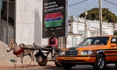 An orange car and a man driving a donkey and cart pass a poster reading, 'Don't let the razorblade be our nation's symbol.'