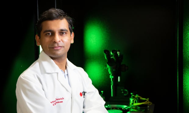 Sumeet Walia is posing for a photo in a lab. He is wearing a lab coat and the lab behind him is dark with a green glow