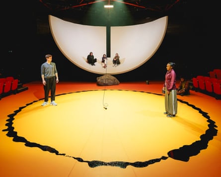 cast members arranged on Alex Lowde’s set that has a shell like shape at the back and a ring of tar on the stage floor.