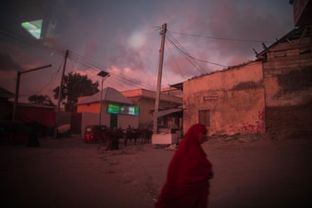 A street scene in Mogadishu. A woman in a red abbaya with her head covered crosses a dusty patch of open ground with decaying single-storey concrete buildings and a few cattle in the background