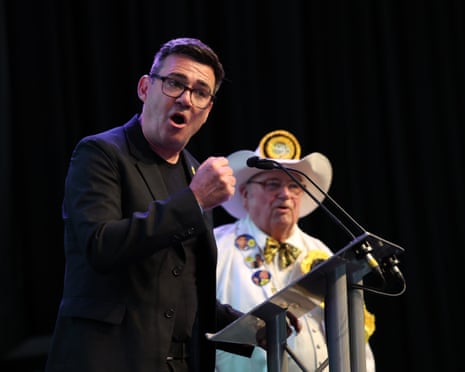 Andy Burnham in glasses and a dark suit speaks passionately at a podium with his fist raised