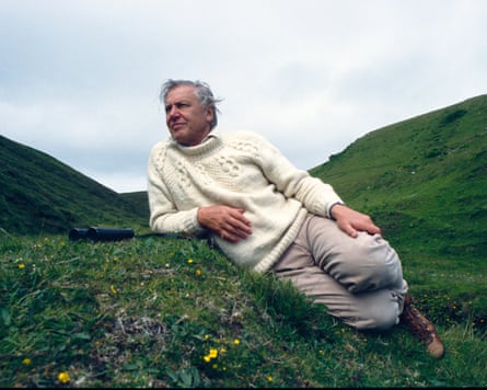 he is reclining on a grassy hillside with a pair of binoculars. He wears a cream knitted woollen jumper and walking boots.