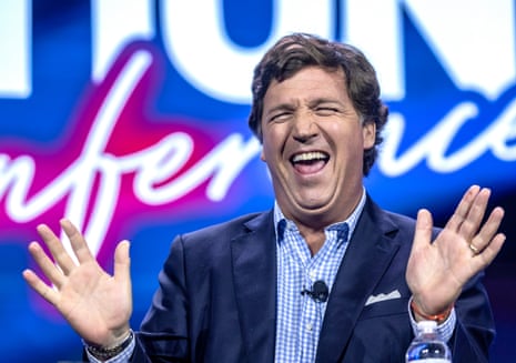 A white man with rakish brown hair, a navy blazer and a blue-and-white gingham shirt with no tie, laughs with his eyes closed as he throws up both palms. He appears to be on a stage, in front of a background of large, unreadable, blue and pink neon letters.
