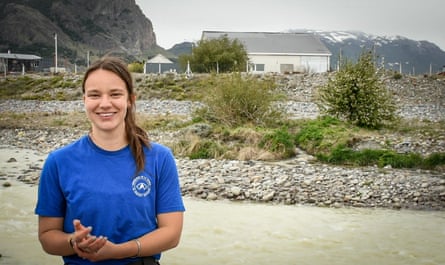 A smiling young woman standing by a river with mountains in the background