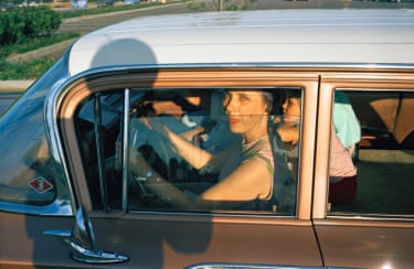 A woman in a pink and green sleeveless dress driving a brown and white-roofed car with a child in the back seat