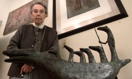 Leonora Carrington, seen here at the age of 84 in her house in Mexico City; she has grey hair tied back into a bun and wears a grey button-front dress and sleeveless jacket over a white polo-neck jumper. She is standing behind one of her sculptures, of a fantastical crocodile-like creature with open jaws and sharp teeth which has five more tall, elongated creatures riding on its back, the tallest holding a long staff. Several of her paintings are seen in the background.