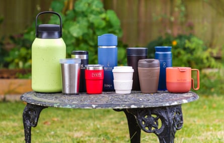 An array of reusable coffee cups and travel mugs on a metal table in a garden.