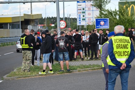 A group of people, mainly young men and some women, stand on the edge of a road near what appears to be border controls; many wear shorts and black hoodies; one man has a T-shirt with a large eagle symbol and Polska on the back. There are people with hi-vis vests on standing by but the scene appears calm and peaceful and no one is holding signs, banners or flags.