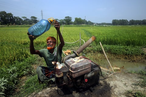 A farmer siphons fuel from a large bottle into a generator sitting by a pond in a paddy field