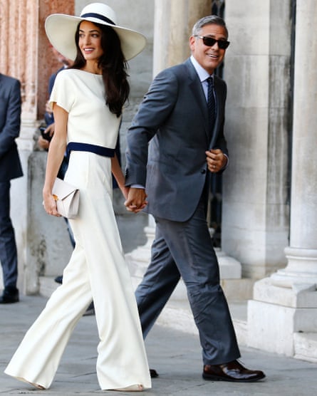 A woman in a white co-ord and floppy hat holds hands with a man in a suit.