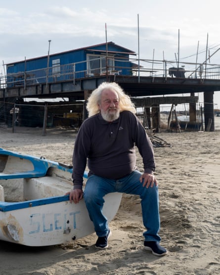 Old man with long greying hair and a beard sitting on a small rowing boat on a beach