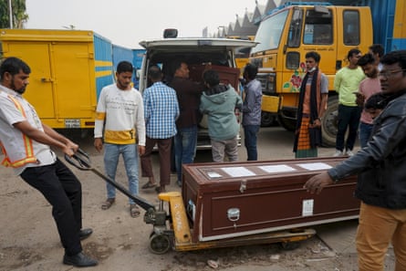A man in a high-vis safety vest pulls a coffin on a trolley as men unload more coffins from a nearby van.