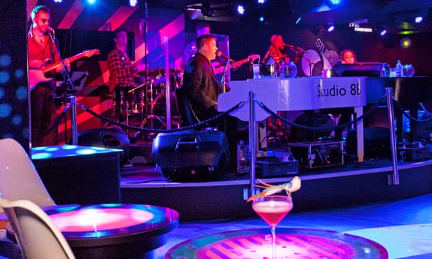 A band playing on a stage with a cocktail on a table in the foreground and a neon wash over the whole scene
