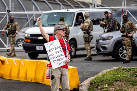 A person holds his fist up while holding a sign that says “The gestapo was just following orders too!” while officers in military gear stand in the background
