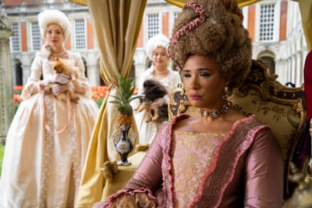 Golda Rosheuvel, with a tall wig and bodiced dress, sits on an embroidered chair, looking serious, with two women in wigs and long dresses behind her
