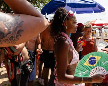 A woman wearing a bikini and holding a fan printed with the Brazilian flag stands on a crowded beach.