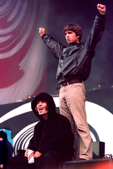 Liam and Noel Gallagher in 1996. Liam sits at the front of the stage wearing a black hoodie and laughing, holding a can of lager, while Noel stands behind him with his arms held up and outstretched.
