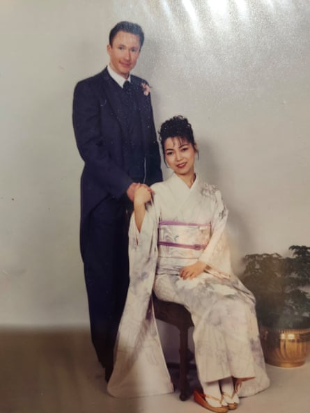 Kerry Cox and Hitomi on their wedding day. Kerry is standing in a navy suit with a pink corsage, and HItomi is seated in a white kimono.