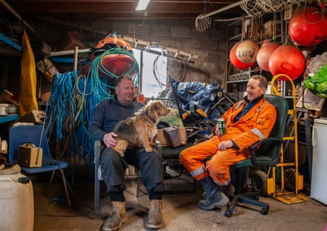 Two mean sit in a shed full of fishing paraphernalia chatting. One has a dog on his lap