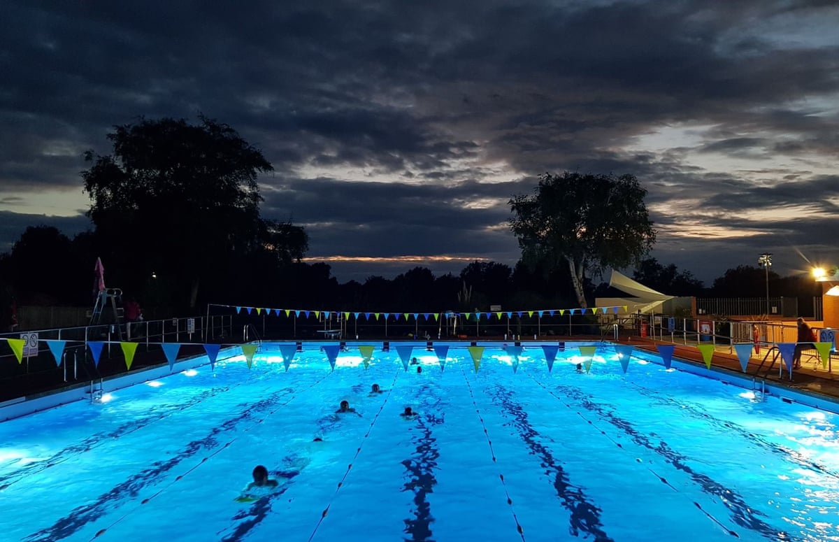 An illuminated outdoor swimming pool against a dark evening sky with silhouetted trees