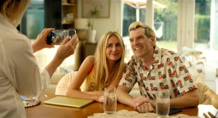 A blond couple pose together cutely at the kitchen table as an unseen third person photographs them on her phone