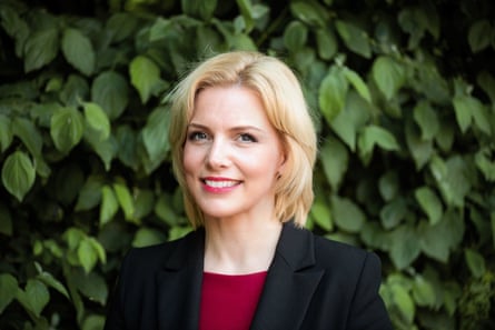she is in her mid-40s and has collar-length blonde hair; she wears a black jacket and dark red top and stands in front of green leaves.