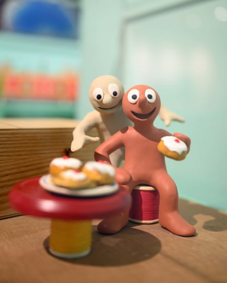 Morph holding a Chelsea bun with Chas behind and several buns on a plate on a table