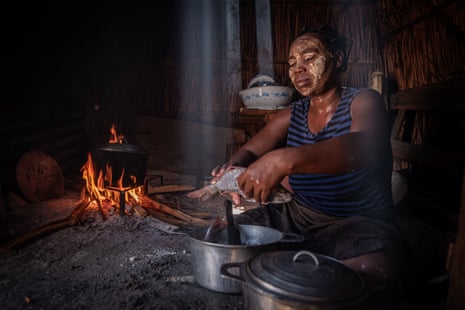 An African woman in a dark hut holds a fish above a cooking pot with a wood fire next to her