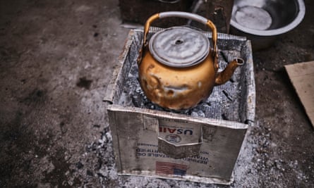 A battered copper-coloured kettle sits on top of a square tin, surrounded by ash.
