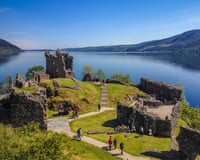 People exploring a ruined castle on a promontory by a loch, with a very blue sky.
