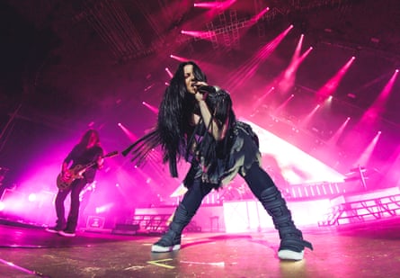 A woman with long black hair wearing all black, in front of a pink light show. A male guitarist is performing behind her