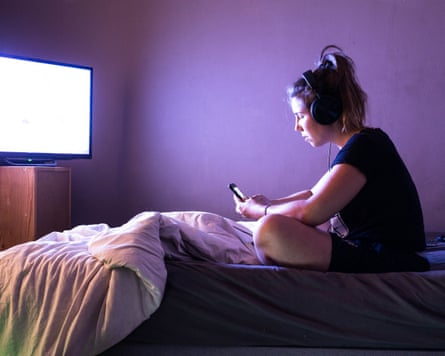 A teenager sits on her bed wearing headphones and looking at her phone. A TV is on in front of her