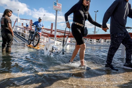 In Newport Beach, people and a dog wade through a flooded street near a ferry ramp