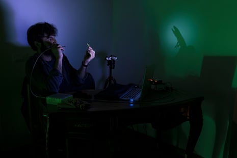 A man holds up a tiny lizard in the dark. It is in a spotlight so its shadow appears on the wall