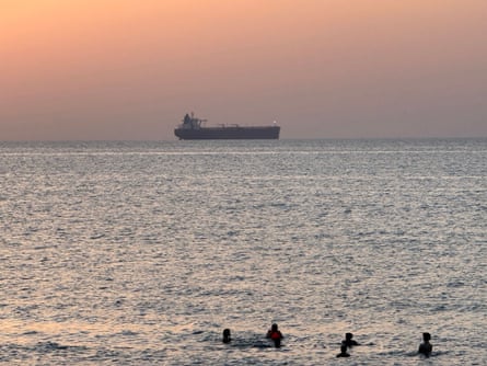A ship is seen on the horizon at sunset with people swimming in the foreground