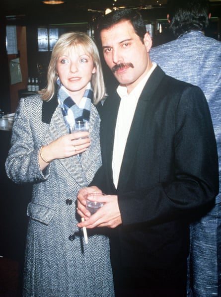 A blond woman in a grey coat and a dark-haired man with a moustache in a dark suit and white shirt, holding a cigarette
