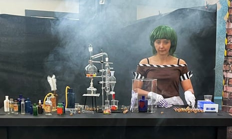 Still image as seen in Channel 5 documentary showing a woman in a green wig playing an Oompa Loompa and looking sad; she is at a work surface with some chemistry equipment, test tubes and bottles