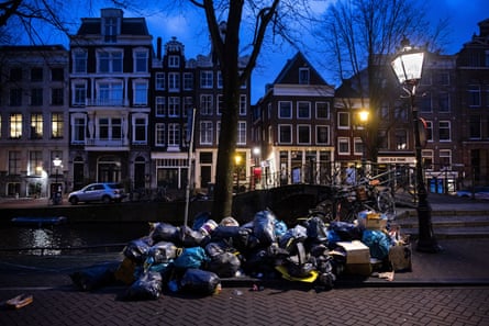 Bags of waste piled up on a street in the centre of Amsterdam by the side of a canal, with traditional brick buildings with gabled roofs in the background.