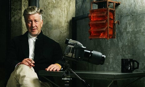 david lynch seated at a table with a camera on a tripod and a cigarette in his hand